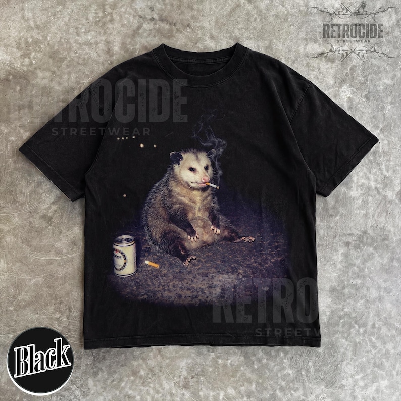 K&ouml;nnte beinhalten: Schwarzes T-Shirt mit einem Aufdruck eines Opossums, das eine Zigarette raucht, mit einer Dose und einer Zigarettenkippe. Auf dem Shirt steht "RETROCIDE STREETWEAR".