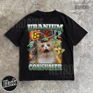 Puede incluir: Una camiseta negra con un estampado gr&aacute;fico de tres gatos rodeados de fuego, rayos verdes y un s&iacute;mbolo de radiaci&oacute;n amarillo. El texto dice URANIUM CONSUMER y 20 BILLION CLORIC. El dise&ntilde;o tiene una est&eacute;tica desgastada de estilo vintage.
