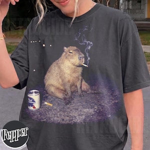 May include: A charcoal gray t-shirt featuring a graphic print of a capybara sitting on the ground with a cigarette in its mouth and a trail of smoke rising above. Next to the capybara is a silver Pabst Blue Ribbon beer can and a cigarette butt.