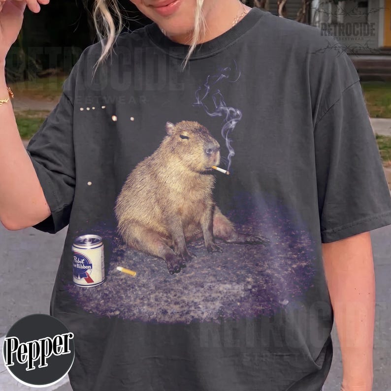 May include: A charcoal gray t-shirt featuring a graphic print of a capybara sitting on the ground with a cigarette in its mouth and a trail of smoke rising above. Next to the capybara is a silver Pabst Blue Ribbon beer can and a cigarette butt.