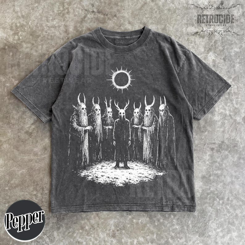 May include: Dark gray t-shirt featuring a white graphic of figures with horned masks and cloaks, standing in a circle under a sun-like symbol. The shirt has a vintage, washed appearance, with the word "RETROCIDE" printed on the top right.