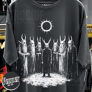May include: Black t-shirt featuring a white graphic of seven figures with horned masks and long robes, standing before a sun-like symbol. The design has a distressed, vintage look, with the word "ROCCIDE" printed at the top.
