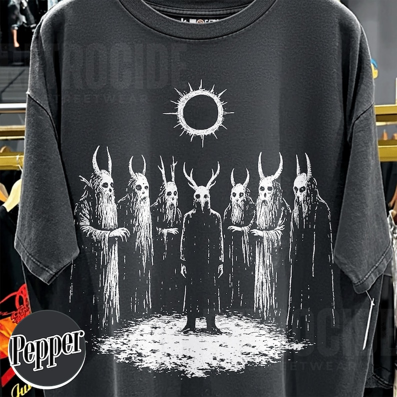 May include: Black t-shirt featuring a white graphic of seven figures with horned masks and long robes, standing before a sun-like symbol. The design has a distressed, vintage look, with the word "ROCCIDE" printed at the top.