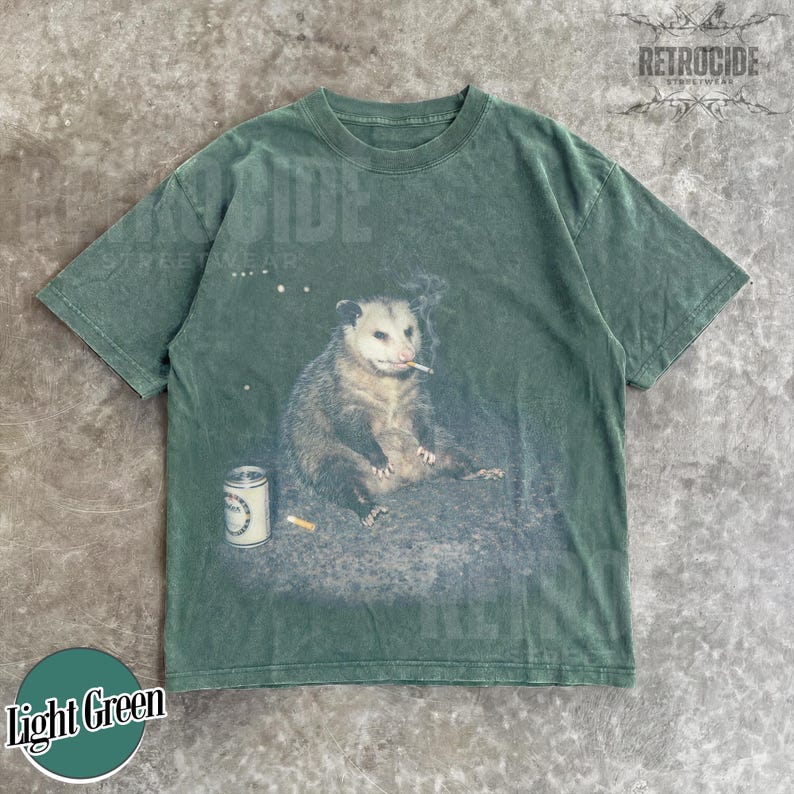 K&ouml;nnte beinhalten: Hellgr&uuml;nes T-Shirt mit einem Aufdruck eines Opossums, das eine Zigarette raucht, mit einer Dose und einem Zigarettenstummel. Das Shirt hat einen Vintage-Look. Der Text "RETROCIDE STREETWEAR" ist oben rechts aufgedruckt.
