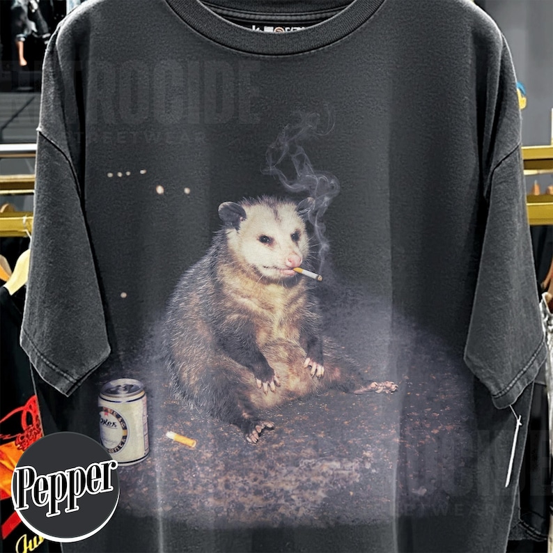 K&ouml;nnte beinhalten: Dunkelgraues T-Shirt mit einem Aufdruck eines Opossums, das eine Zigarette raucht, mit einer Bierdose und einem Zigarettenstummel. Das Wort "ROCCIDE" ist oben aufgedruckt. Streetwear.