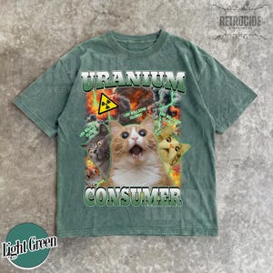 Puede incluir: Una camiseta verde claro con un estampado gr&aacute;fico de tres gatos sobre un fondo de fuego y rayos. El texto dice "URANIUM CONSUMER" y "20 BILLION CLORIC" con un s&iacute;mbolo amarillo de advertencia de radiaci&oacute;n sobre el gato central.