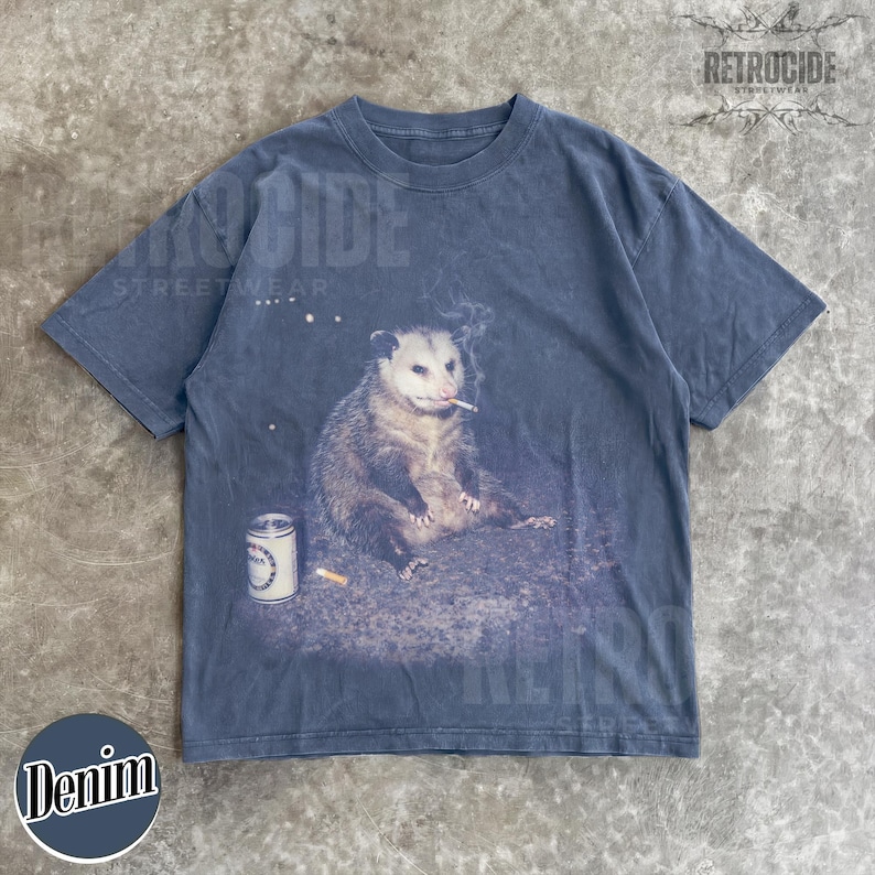 K&ouml;nnte beinhalten: Denimblaues T-Shirt mit einer Grafik eines Opossums, das eine Zigarette raucht, mit einer Dose und einem Zigarettenstummel. Das Shirt hat den Text "RETROCIDE STREETWEAR" und "Denim".
