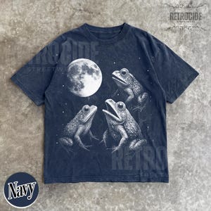 May include: Navy blue t-shirt featuring a detailed illustration of three frogs under a full moon. The graphic is in black and white, with the word "Navy" in a circle at the bottom. The shirt has a vintage, distressed look.