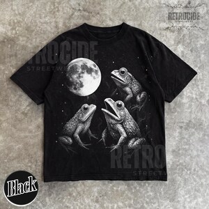 May include: Black t-shirt featuring a detailed illustration of three frogs and a full moon. The design is in grayscale, with the frogs positioned around the moon. The shirt has the text "RETROCIDE STREETWEAR" printed on it.
