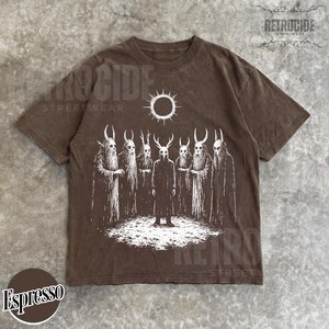 May include: Espresso-colored t-shirt featuring a white graphic of figures with horned masks and cloaks, standing beneath a sun symbol. The shirt has a vintage, washed look, with the word "RETROCIDE" printed on the top right.