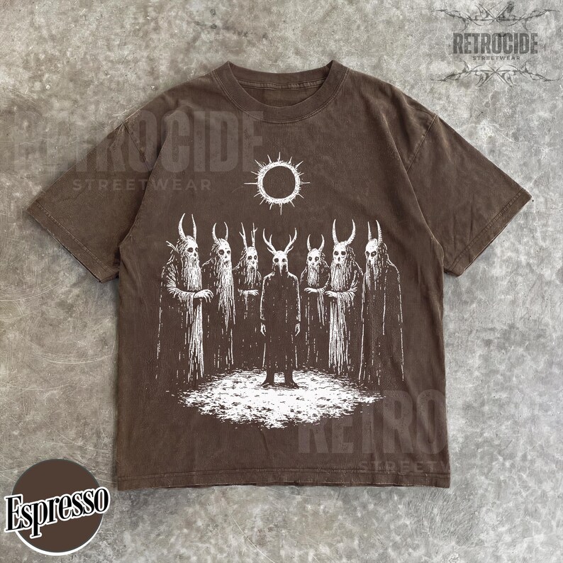 May include: Espresso-colored t-shirt featuring a white graphic of figures with horned masks and cloaks, standing beneath a sun symbol. The shirt has a vintage, washed look, with the word "RETROCIDE" printed on the top right.