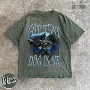 Peut inclure: T-shirt vert mousse avec un motif de loup-garou et le texte "GOT THAT DOG IN ME". Le design pr&eacute;sente trois loups-garous dans des tons bleus et gris. Le t-shirt a un aspect vintage et vieilli.