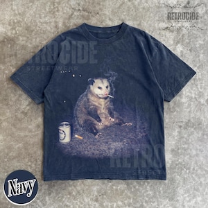 K&ouml;nnte beinhalten: Marineblaues T-Shirt mit einem Motiv eines Opossums, das eine Zigarette raucht, mit einer Dose und einem Zigarettenstummel. Das Shirt hat einen Vintage-Look. Das Wort "RETROCIDE" ist oben auf dem Shirt aufgedruckt.