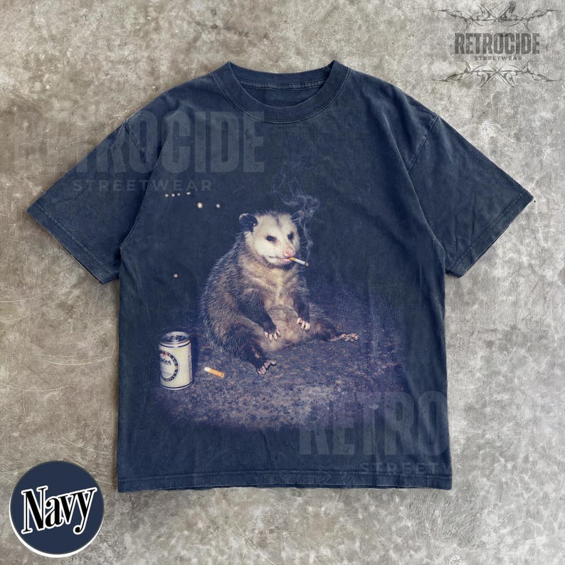 K&ouml;nnte beinhalten: Marineblaues T-Shirt mit einem Motiv eines Opossums, das eine Zigarette raucht, mit einer Dose und einem Zigarettenstummel. Das Shirt hat einen Vintage-Look. Das Wort "RETROCIDE" ist oben auf dem Shirt aufgedruckt.