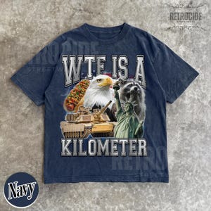 May include: Navy blue t-shirt with a graphic design featuring a hot dog, bald eagle, raccoon, tank, and the Statue of Liberty. The text "WTF IS A KILOMETER" is printed across the front. The shirt has a vintage, distressed look.