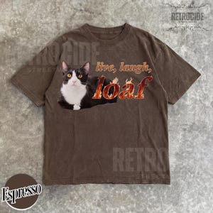Puede incluir: Una camiseta marr&oacute;n de manga corta con un gr&aacute;fico de un gato blanco y negro tumbado. El texto dice "live, laugh, loaf" con la palabra "loaf" estilizada en un patr&oacute;n de llamas de color naranja y amarillo. La camiseta tiene cuello redondo.
