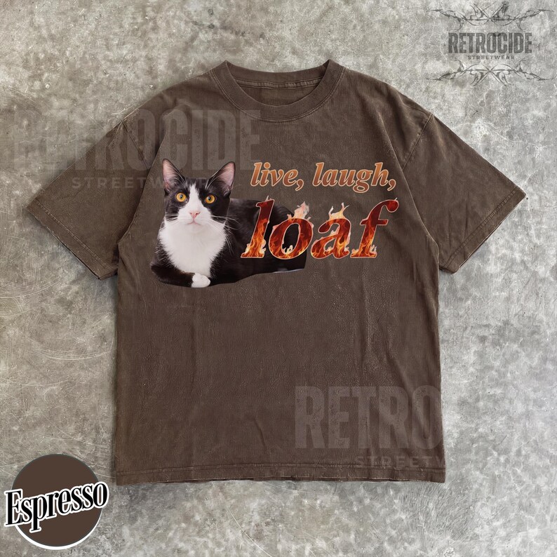 Puede incluir: Una camiseta marr&oacute;n de manga corta con un gr&aacute;fico de un gato blanco y negro tumbado. El texto dice "live, laugh, loaf" con la palabra "loaf" estilizada en un patr&oacute;n de llamas de color naranja y amarillo. La camiseta tiene cuello redondo.