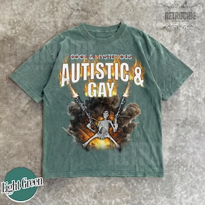 May include: Light green t-shirt with the text "COOL & MYSTERIOUS AUTISTIC & GAY" in a bold, stylized font. The graphic features a skeleton figure holding two guns with flames and smoke. The shirt has a vintage, worn look.