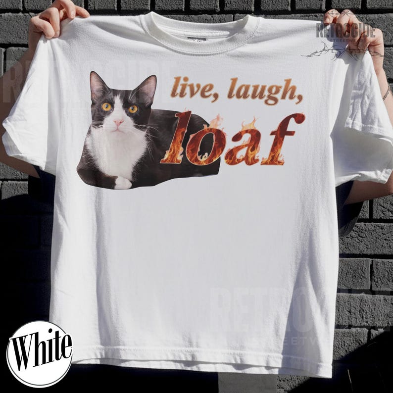 Puede incluir: Una camiseta blanca con un gr&aacute;fico de un gato blanco y negro en posici&oacute;n de hogaza. El texto "live, laugh," est&aacute; impreso en marr&oacute;n sobre la palabra "loaf," estilizada con un patr&oacute;n de llamas naranjas y amarillas sobre las letras.