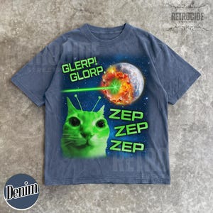 May include: Denim blue t-shirt with a green alien cat graphic. The graphic includes the words "GLERP! GLORP." and "ZEP ZEP ZEP" with a planet being zapped by a laser beam. The shirt has a vintage streetwear aesthetic.