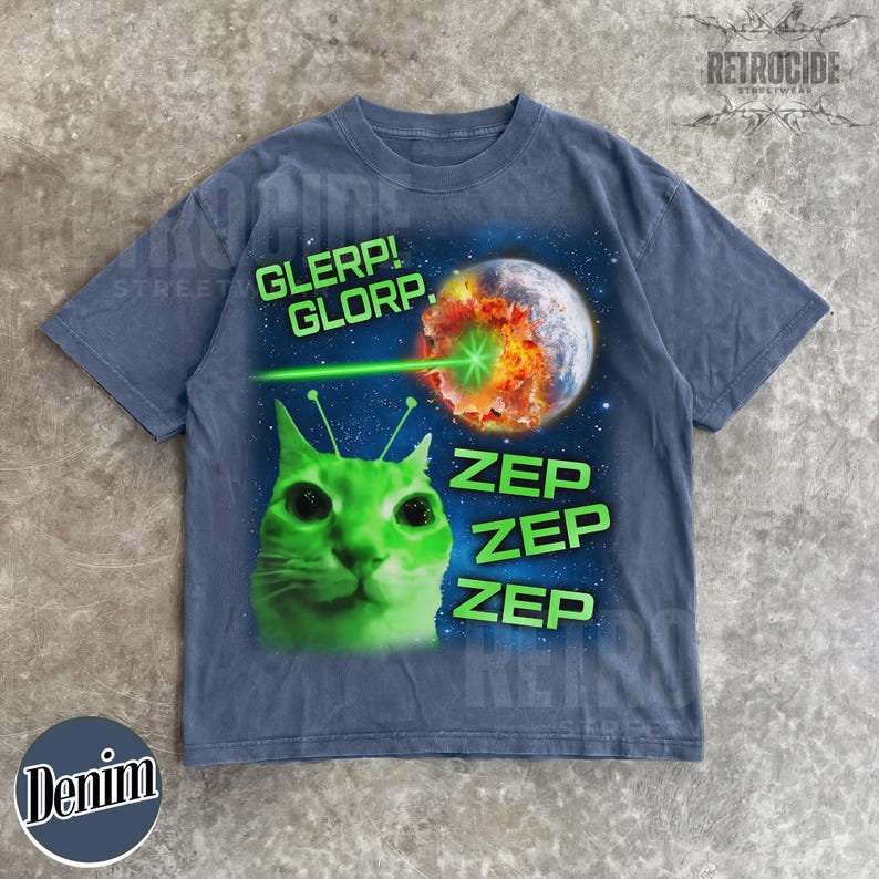 May include: Denim blue t-shirt with a green alien cat graphic. The graphic includes the words "GLERP! GLORP." and "ZEP ZEP ZEP" with a planet being zapped by a laser beam. The shirt has a vintage streetwear aesthetic.