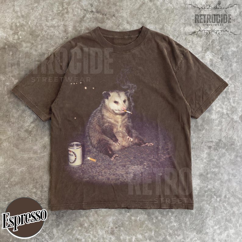 K&ouml;nnte beinhalten: Espressofarbenes T-Shirt mit einer Grafik eines Opossums, das eine Zigarette raucht, mit einer Dose und einem Zigarettenstummel in der N&auml;he. Das Shirt tr&auml;gt den Text "RETROCIDE STREETWEAR".