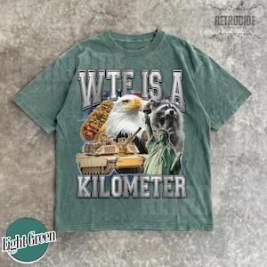 May include: Light green t-shirt with a graphic design featuring a hot dog, bald eagle, raccoon, tank, and the Statue of Liberty. The text reads "WTF IS A KILOMETER". The shirt has a vintage, distressed look.