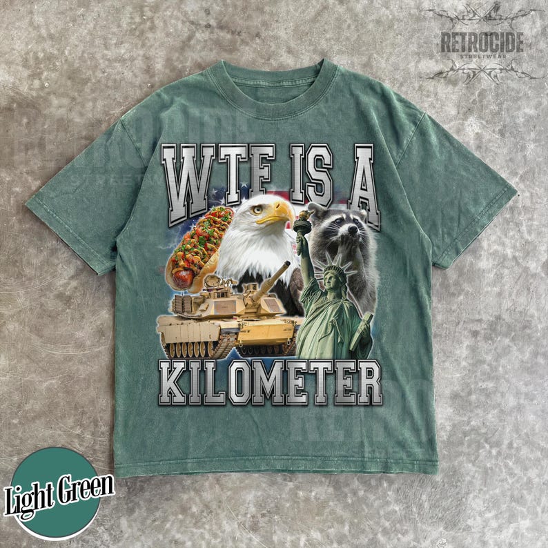 May include: Light green t-shirt with a graphic design featuring a hot dog, bald eagle, raccoon, tank, and the Statue of Liberty. The text reads "WTF IS A KILOMETER". The shirt has a vintage, distressed look.