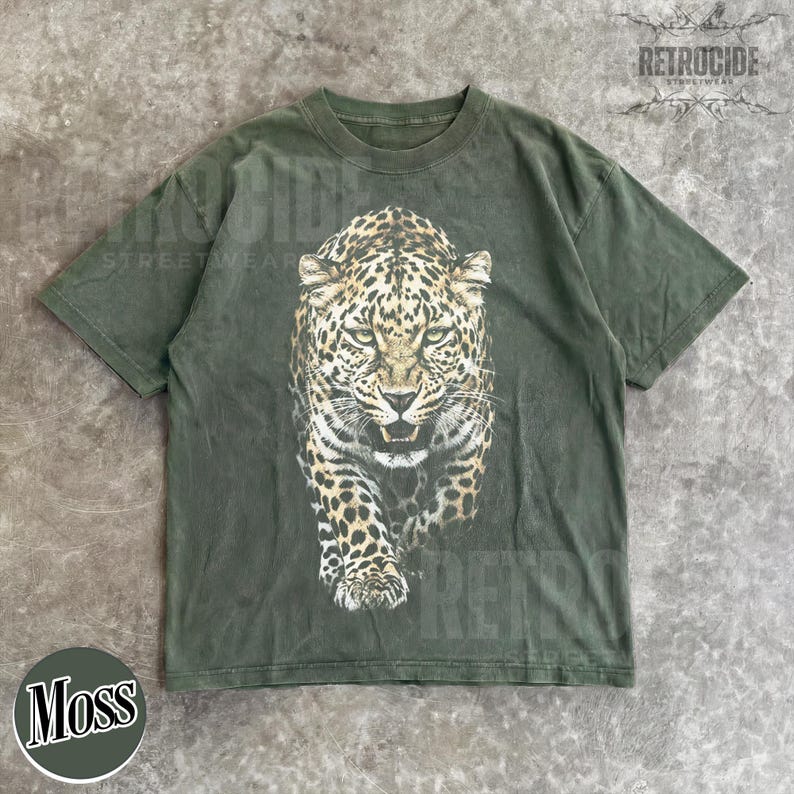 May include: Green t-shirt featuring a detailed leopard graphic. The leopard is depicted in shades of brown, black, and white, with a fierce expression. The shirt has the word "RETROCIDE" printed on it. The shirt is labeled "Moss".