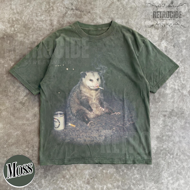 K&ouml;nnte beinhalten: Moosgr&uuml;nes T-Shirt mit einem Aufdruck eines Opossums, das eine Zigarette raucht, mit einer Bierdose und einem Zigarettenstummel. Das Shirt hat einen Vintage-Look mit dem Text "RETROCIDE STREETWEAR" oben rechts.
