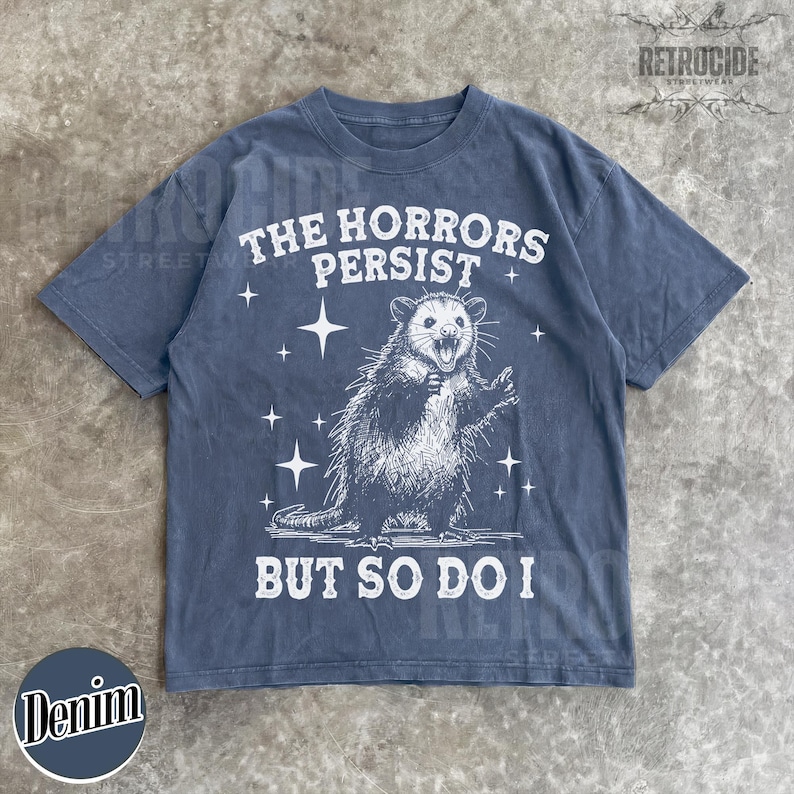 May include: Denim blue t-shirt with a distressed graphic of an opossum and the text "THE HORRORS PERSIST BUT SO DO I". The shirt has a crew neck and short sleeves. The brand name "RETROCIDE" is printed on the top right.