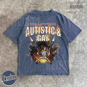 May include: Denim blue t-shirt with the text "COOL & MYSTERIOUS" above "AUTISTIC & GAY" in a bold, white font. A skeleton figure is depicted holding two guns with flames and smoke. The shirt has a vintage streetwear aesthetic.