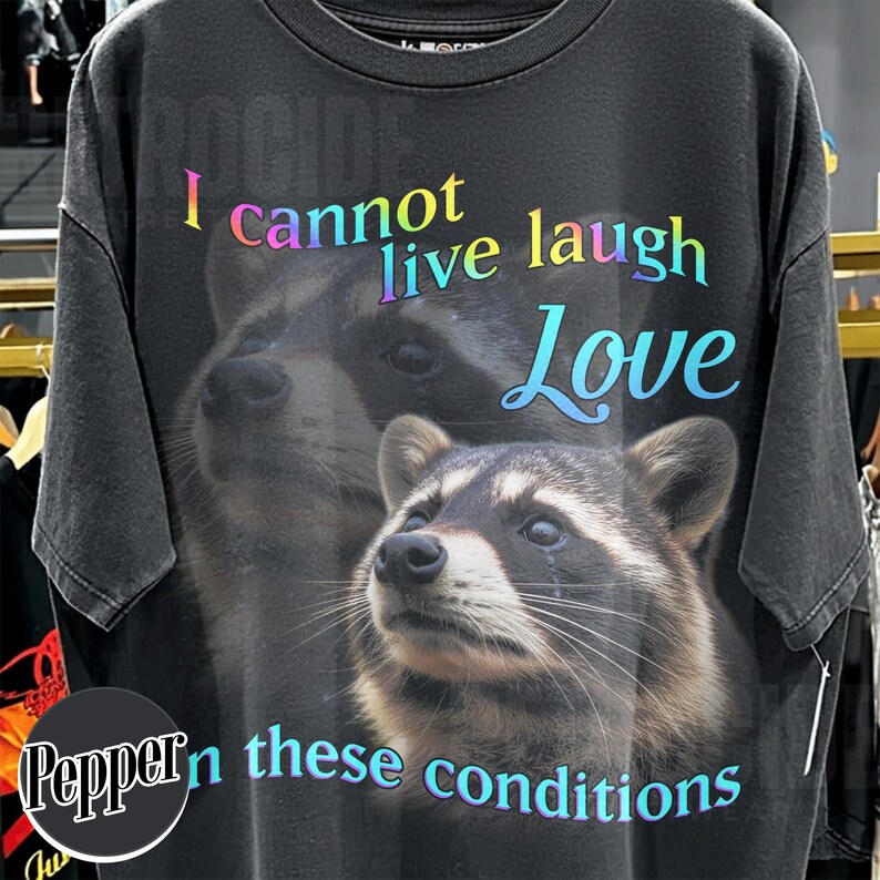 Puede incluir: Una camiseta gris carb&oacute;n con un gr&aacute;fico grande de dos mapaches. El texto "I cannot live laugh Love" y "in these conditions" est&aacute; impreso en una fuente con degradado de arco&iacute;ris en la parte delantera, sobre la imagen de los mapaches.