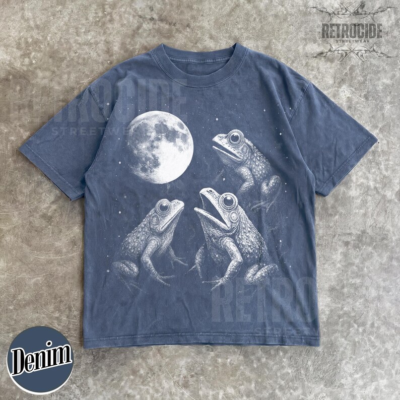 May include: Denim blue t-shirt featuring a graphic of three frogs under a full moon. The frogs are detailed in shades of gray and white, with the moon in the background. The shirt has a crew neck and short sleeves.
