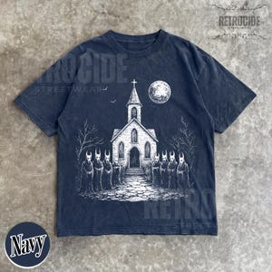 Op de afbeelding: Een marineblauw t-shirt met korte mouwen en een witte grafische print van een kerk met een toren, een volle maan en een groep figuren in capuchons met gehoornde maskers voor het gebouw. De tekst RETROCIDE STREETWEAR is zichtbaar.