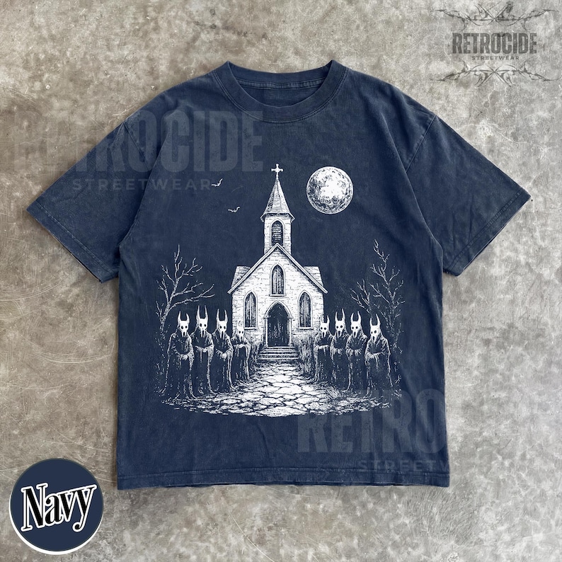 Op de afbeelding: Een marineblauw t-shirt met korte mouwen en een witte grafische print van een kerk met een toren, een volle maan en een groep figuren in capuchons met gehoornde maskers voor het gebouw. De tekst RETROCIDE STREETWEAR is zichtbaar.