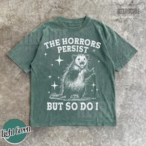 May include: Light green t-shirt with a distressed graphic of an opossum and the text "THE HORRORS PERSIST BUT SO DO I." The shirt has a crew neck and short sleeves. The brand name "RETROCIDE" is printed at the top right.