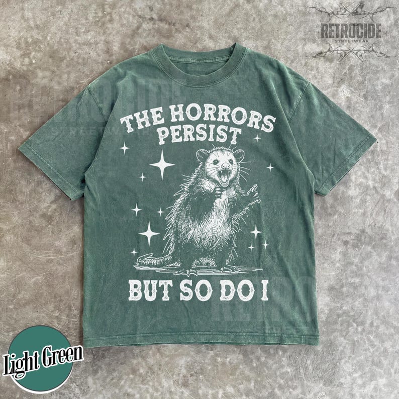 May include: Light green t-shirt with a distressed graphic of an opossum and the text "THE HORRORS PERSIST BUT SO DO I." The shirt has a crew neck and short sleeves. The brand name "RETROCIDE" is printed at the top right.