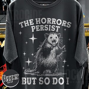 May include: Black t-shirt with a distressed graphic of an opossum with the text "THE HORRORS PERSIST BUT SO DO I". The opossum is illustrated in a detailed, vintage style, surrounded by star shapes.