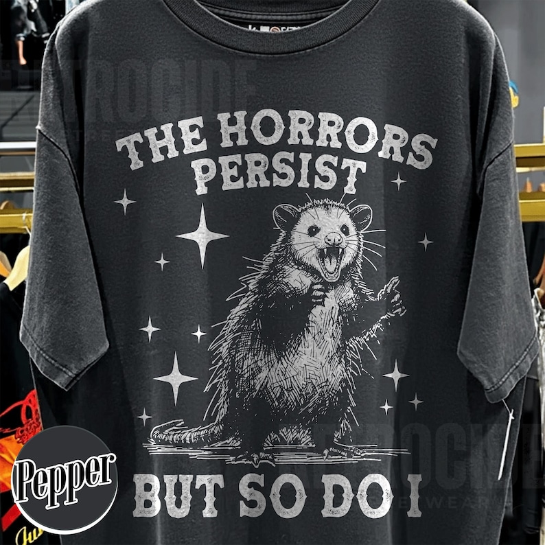 May include: Black t-shirt with a distressed graphic of an opossum with the text "THE HORRORS PERSIST BUT SO DO I". The opossum is illustrated in a detailed, vintage style, surrounded by star shapes.
