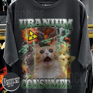 Puede incluir: Una camiseta gris carb&oacute;n con un estampado gr&aacute;fico que incluye el texto URANIUM CONSUMER. El dise&ntilde;o muestra tres gatos, un s&iacute;mbolo amarillo de advertencia de radiaci&oacute;n, rayos verdes y el texto +20 BILLIOW CLORIC sobre un fondo de fuego.