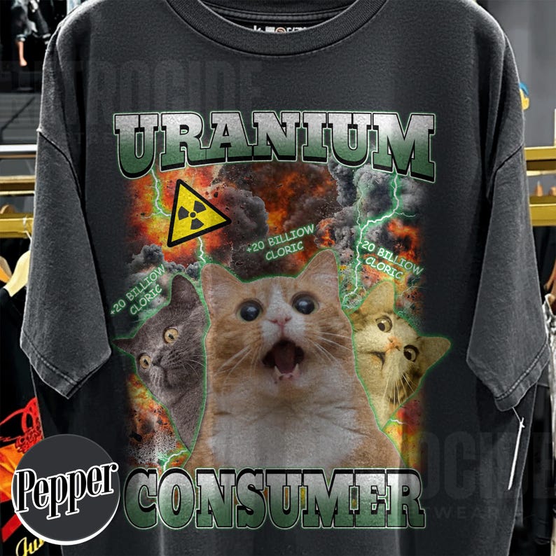 Puede incluir: Una camiseta gris carb&oacute;n con un estampado gr&aacute;fico que incluye el texto URANIUM CONSUMER. El dise&ntilde;o muestra tres gatos, un s&iacute;mbolo amarillo de advertencia de radiaci&oacute;n, rayos verdes y el texto +20 BILLIOW CLORIC sobre un fondo de fuego.
