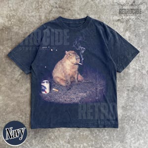 May include: A navy blue short-sleeve t-shirt featuring a graphic print of a capybara sitting on the ground with a lit cigarette in its mouth, smoke rising above, and a Pabst Blue Ribbon beer can placed on the ground next to the animal.