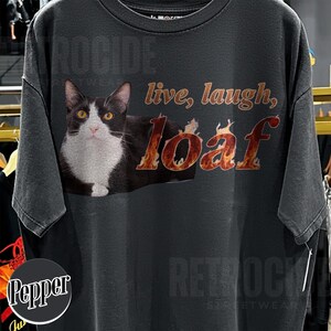 Puede incluir: Una camiseta gris carb&oacute;n de manga corta con el gr&aacute;fico de un gato blanco y negro. El texto "live, laugh," est&aacute; impreso en marr&oacute;n y la palabra "loaf" se muestra con un patr&oacute;n de llamas de color naranja y amarillo en el pecho.