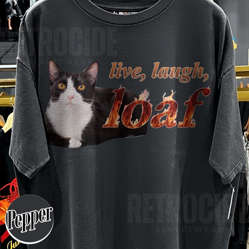 Puede incluir: Una camiseta gris carb&oacute;n de manga corta con el gr&aacute;fico de un gato blanco y negro. El texto "live, laugh," est&aacute; impreso en marr&oacute;n y la palabra "loaf" se muestra con un patr&oacute;n de llamas de color naranja y amarillo en el pecho.