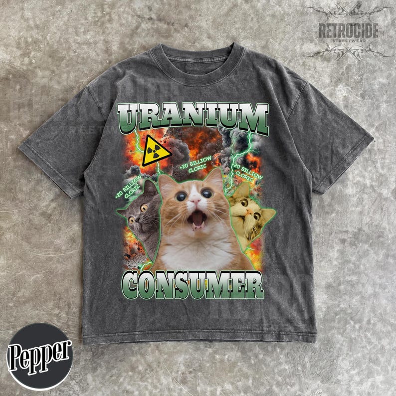 May include: A charcoal gray short-sleeve t-shirt featuring a graphic print of three cats against a fiery explosion background. The text reads URANIUM CONSUMER with a yellow radiation symbol and the phrase 20 BILLION CLORIC repeated twice.