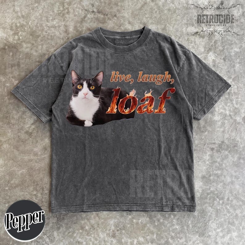 Puede incluir: Una camiseta de manga corta gris carb&oacute;n con un gr&aacute;fico de un gato blanco y negro tumbado. Junto al gato, el texto dice "live, laugh, loaf" con la palabra "loaf" estilizada en un dise&ntilde;o de patr&oacute;n de llamas de color naranja y amarillo.