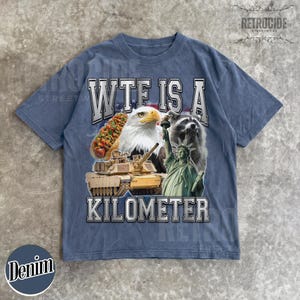 May include: Denim blue t-shirt with a graphic design featuring a hot dog, bald eagle, raccoon, tank, and the Statue of Liberty. The text on the shirt reads "WTF IS A KILOMETER". The shirt has a vintage, distressed look.