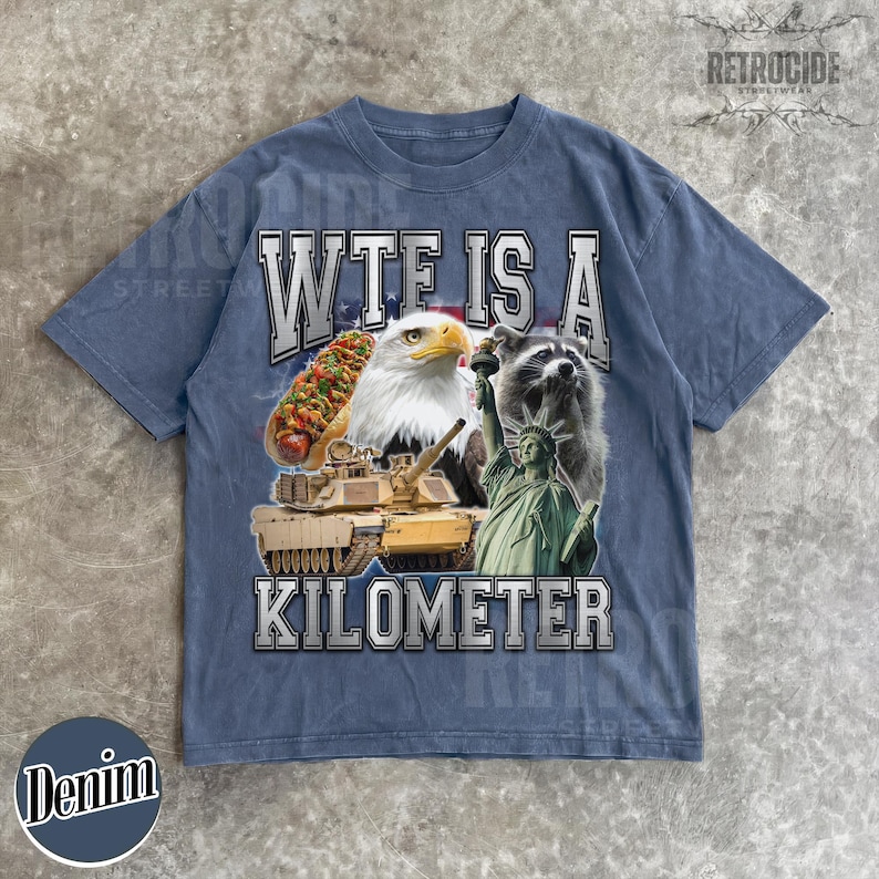 May include: Denim blue t-shirt with a graphic design featuring a hot dog, bald eagle, raccoon, tank, and the Statue of Liberty. The text on the shirt reads "WTF IS A KILOMETER". The shirt has a vintage, distressed look.