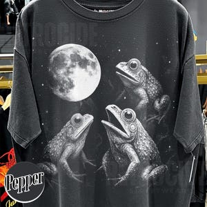 May include: Dark gray t-shirt featuring a detailed illustration of three frogs and a full moon against a starry night sky. The design is in black and white, creating a vintage aesthetic. The word "ROCKS" is printed at the top.